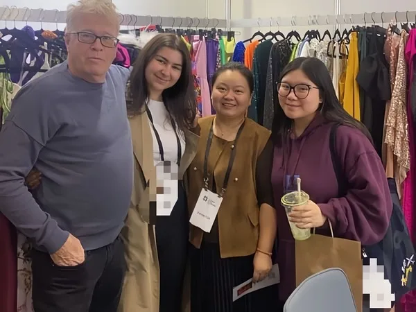 Four people stand together in front of a colorful clothing rack, all smiles as they prepare to attend the Melbourne fashion show. Three of them wear lanyards with nametags, and one holds a drink cup, adding to the friendly atmosphere.